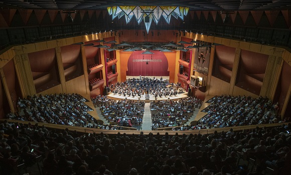 Choir and orchestra performing in front of a large window with a sea view.
