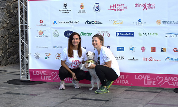 Two smiling women pose with a dog in front of a sponsor banner.